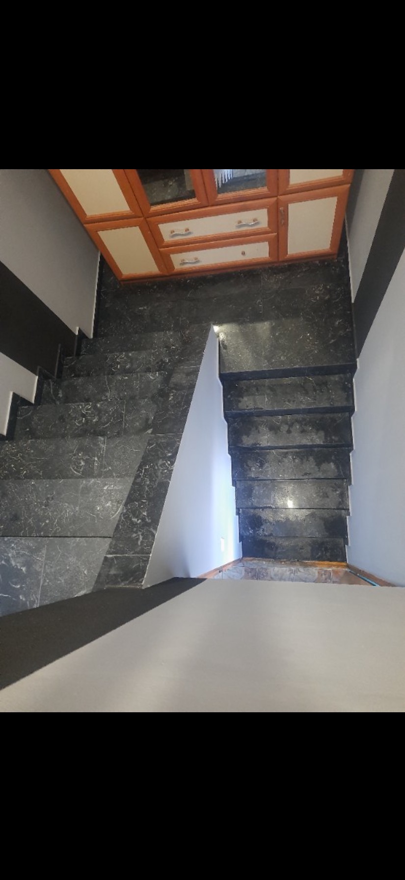 Schody wyłożone ciemnym gresem z wyraźnym wzorem marmuru, prowadzące w dół, z szafką wiszącą nad nimi i pomalowaną ścianą po prawej stronie.