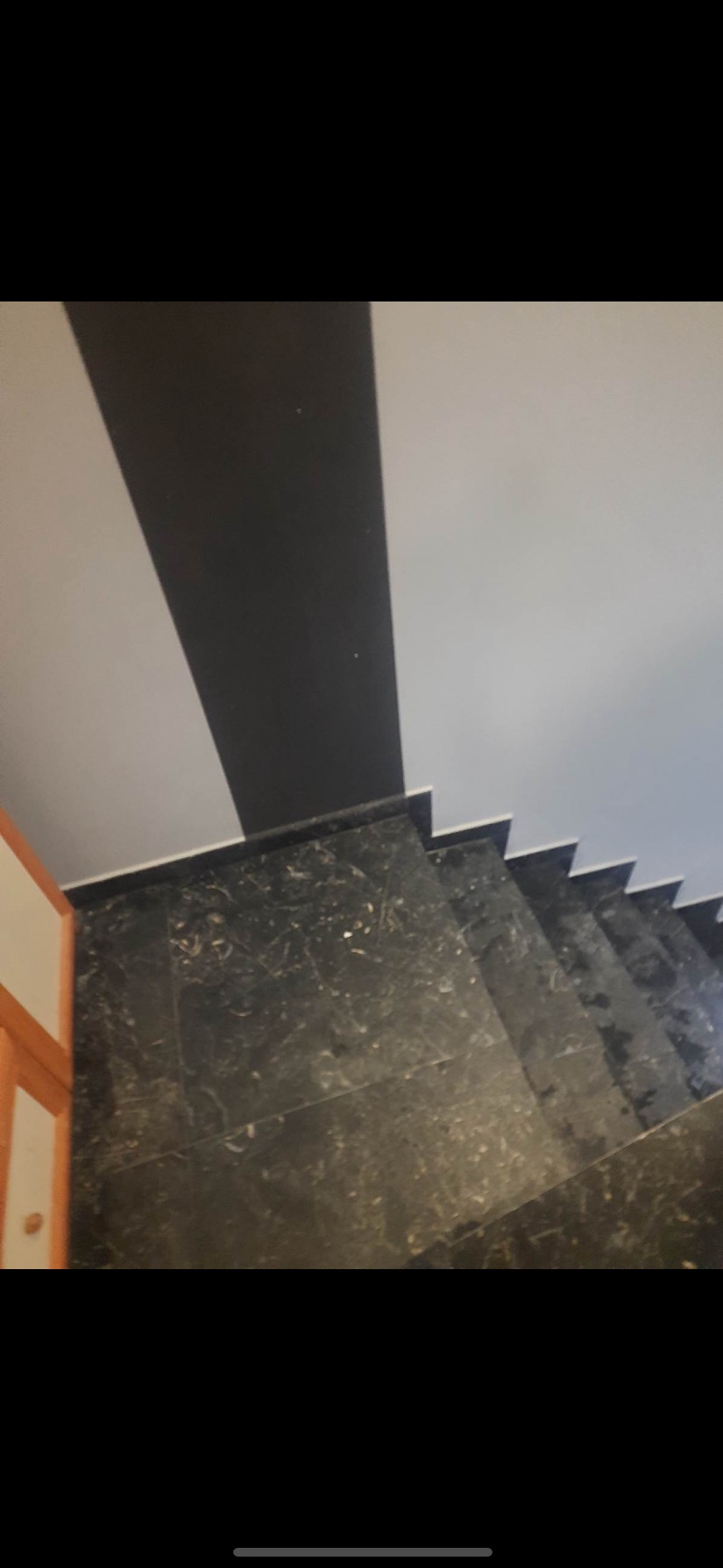 Widok z góry na schody wyłożone ciemnym marmurem z widocznymi żyłkami, obok fragment ściany pomalowanej w szerokie, naprzemienne pasy szarości i bieli, oraz fragment drewnianej framugi drzwi.