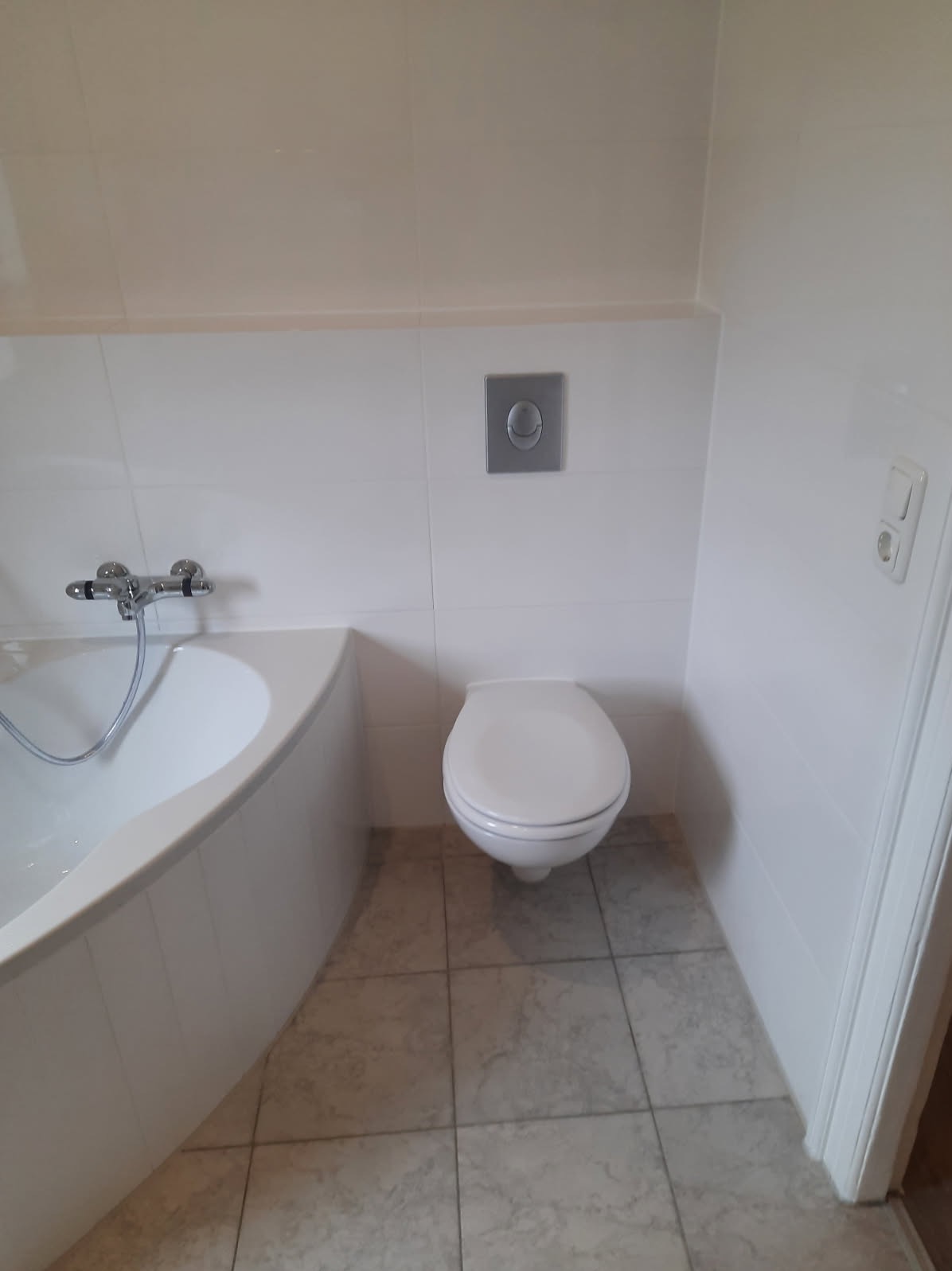 Biała łazienka z wanną narożną, toaletą podtynkową i beżowymi płytkami podłogowymi, sfotografowana z perspektywy lekko z góry.