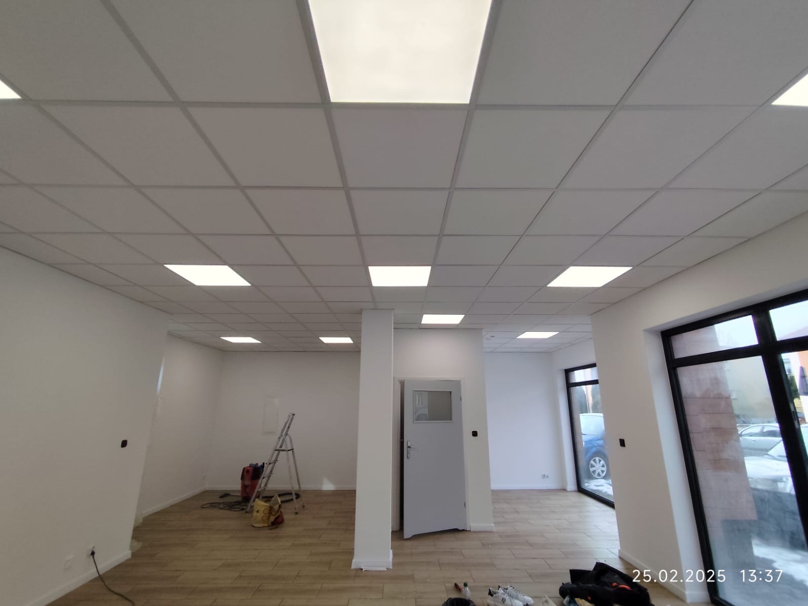 Wnętrze biura po remoncie z podwieszanym sufitem kasetonowym z oświetleniem LED, świeżo pomalowane ściany, drewnopodobna podłoga, widoczny sprzęt malarski i drabina.