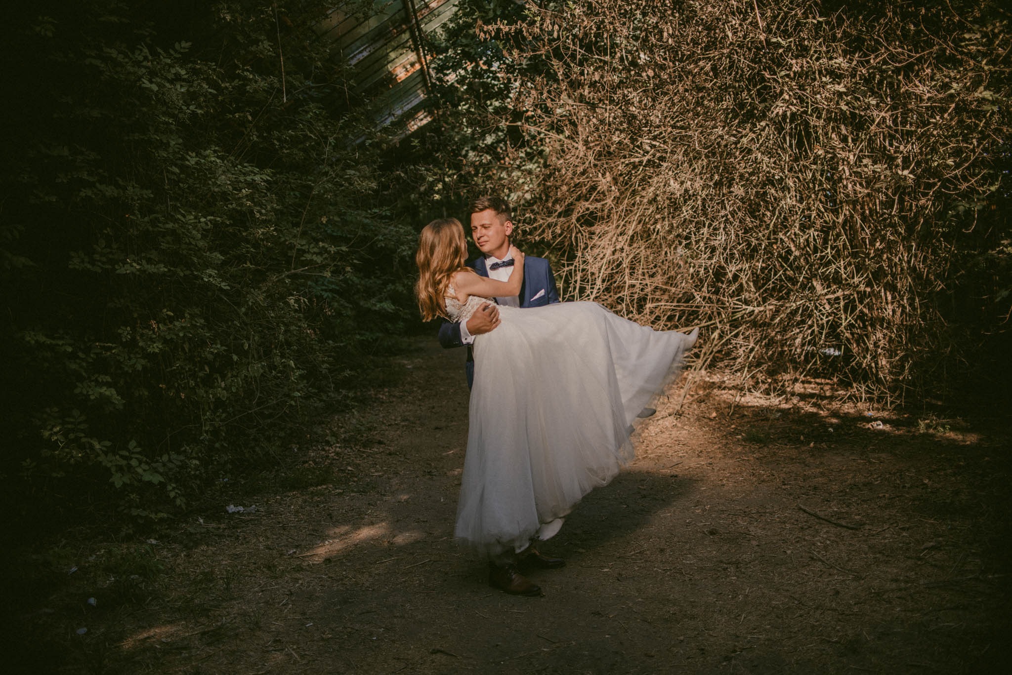 Uśmiechnięty pan młody w granatowej marynarce i muszce trzyma na rękach pannę młodą w białej sukni ślubnej, na tle zarośli i metalowej konstrukcji szklarni.