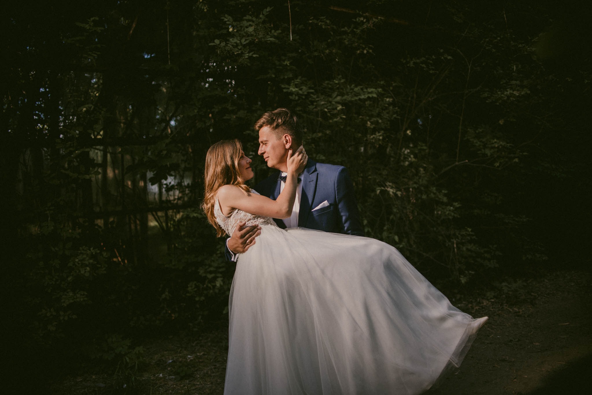 Uśmiechnięta panna młoda w koronkowej sukni ślubnej, niesiona na rękach przez pana młodego w niebieskim garniturze na tle zieleni.