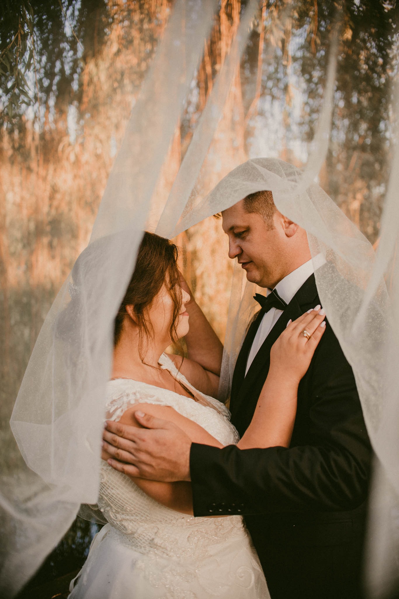 Para młoda w objęciach pod delikatnym, zwiewnym welonem na tle rozmytej zieleni i złocistego światła.