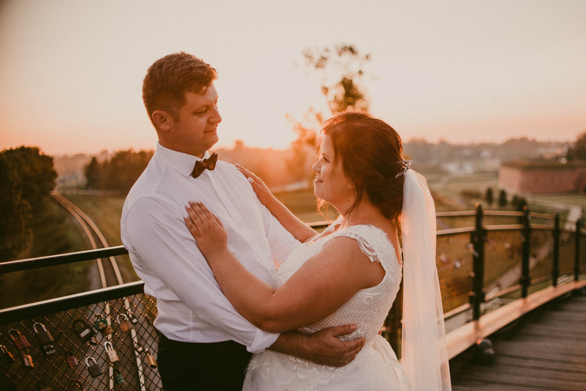 Para młoda w objęciach na drewnianym moście o zachodzie słońca, z widokiem na tor kolejowy i twierdzę w tle, kłódki miłości na barierkach mostu.