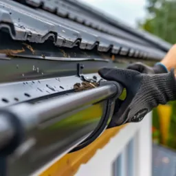 Pracownik w rękawicach usuwa zanieczyszczenia z czarnej rynny zamontowanej na dachu budynku.