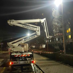 WEZA Mirosław Sitko - Biały podnośnik koszowy na chodniku oświetlonym latarnią, w tle budynek z oknami, na latarni zawieszone dekoracje świąteczne.