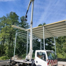 WEZA Mirosław Sitko - Montaż płyty warstwowej na stalowej konstrukcji z użyciem podnośnika koszowego. Widoczny fragment lasu w tle i niebieskie niebo.