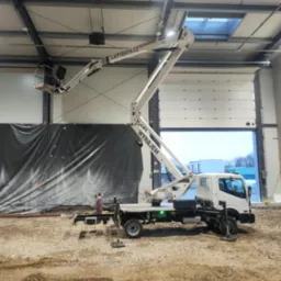 Podnośnik koszowy z pracownikiem w koszu, na tle hali z otwartą bramą. Widoczny fragment elewacji i plac budowy. Na ramieniu podnośnika numer telefonu.