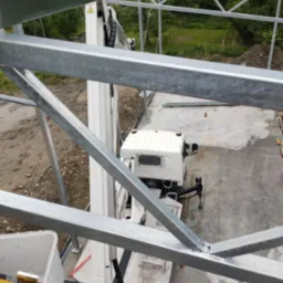 Widok z góry na konstrukcję metalową w trakcie montażu, z użyciem podnośnika koszowego. Widoczne elementy konstrukcji, podnośnik oraz otoczenie placu budowy.