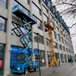 WEZA Mirosław Sitko - Dwa podnośniki koszowe: niebieski i żółty, ustawione przy szarym budynku z licznymi oknami. Jeden pracownik na żółtym podnośniku. Gałęzie drzew na pierwszym planie.