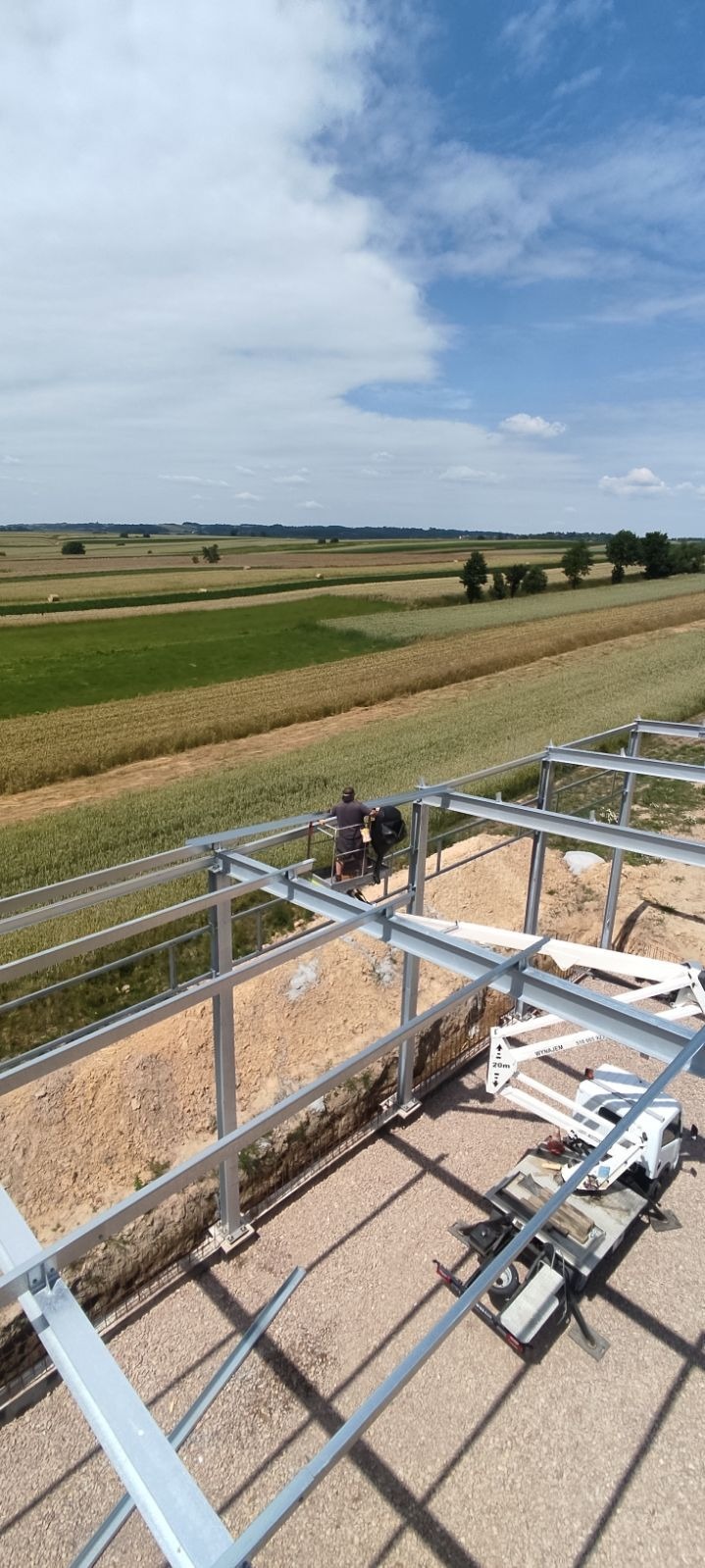Pracownicy na podnośniku koszowym montują konstrukcję stalową budynku gospodarczego na tle pól uprawnych i błękitnego nieba z chmurami.