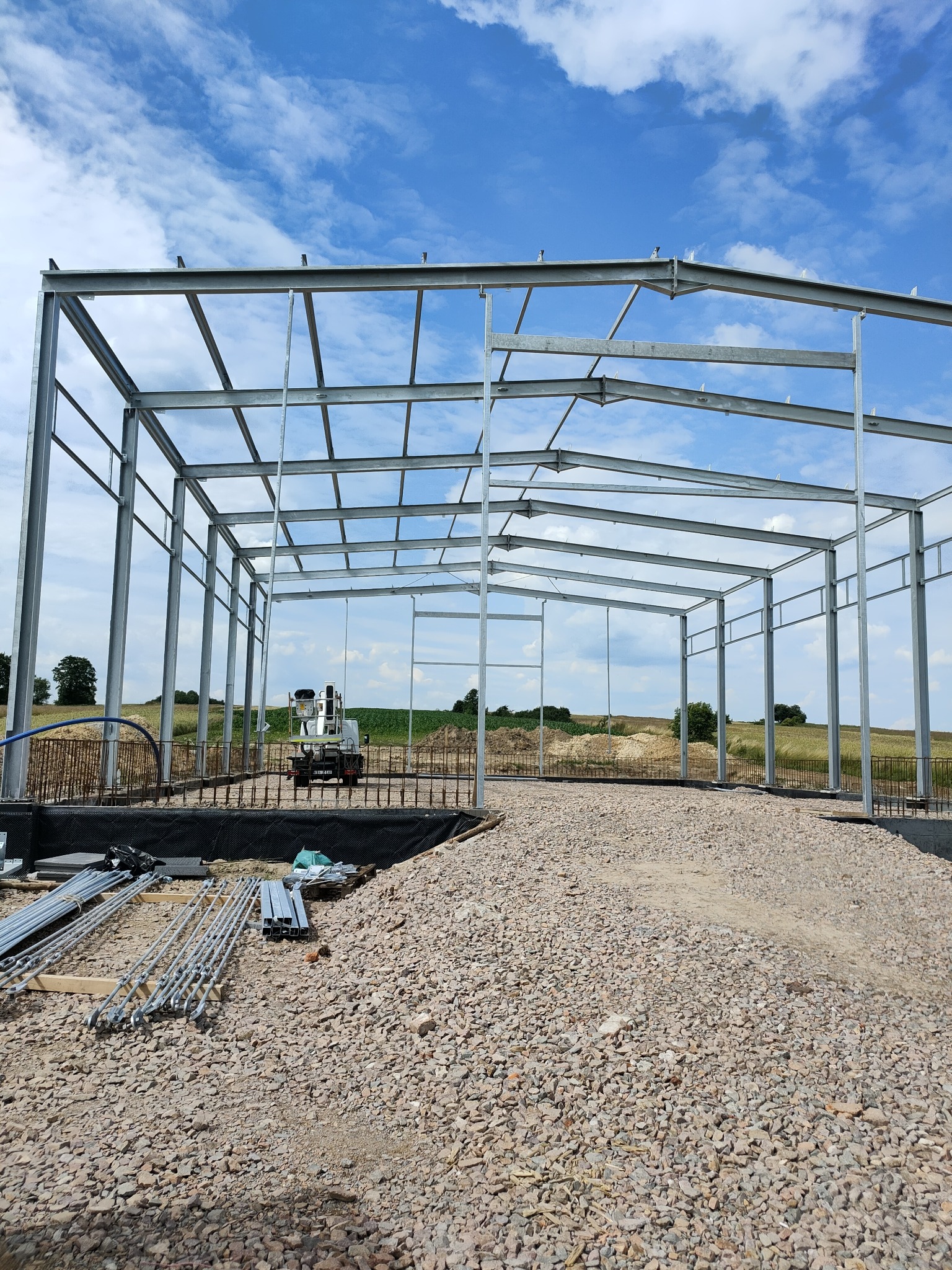 Stalowa konstrukcja szkieletowa budynku w trakcie montażu, widoczny podnośnik koszowy w tle oraz przygotowane elementy konstrukcyjne na żwirowym podłożu.