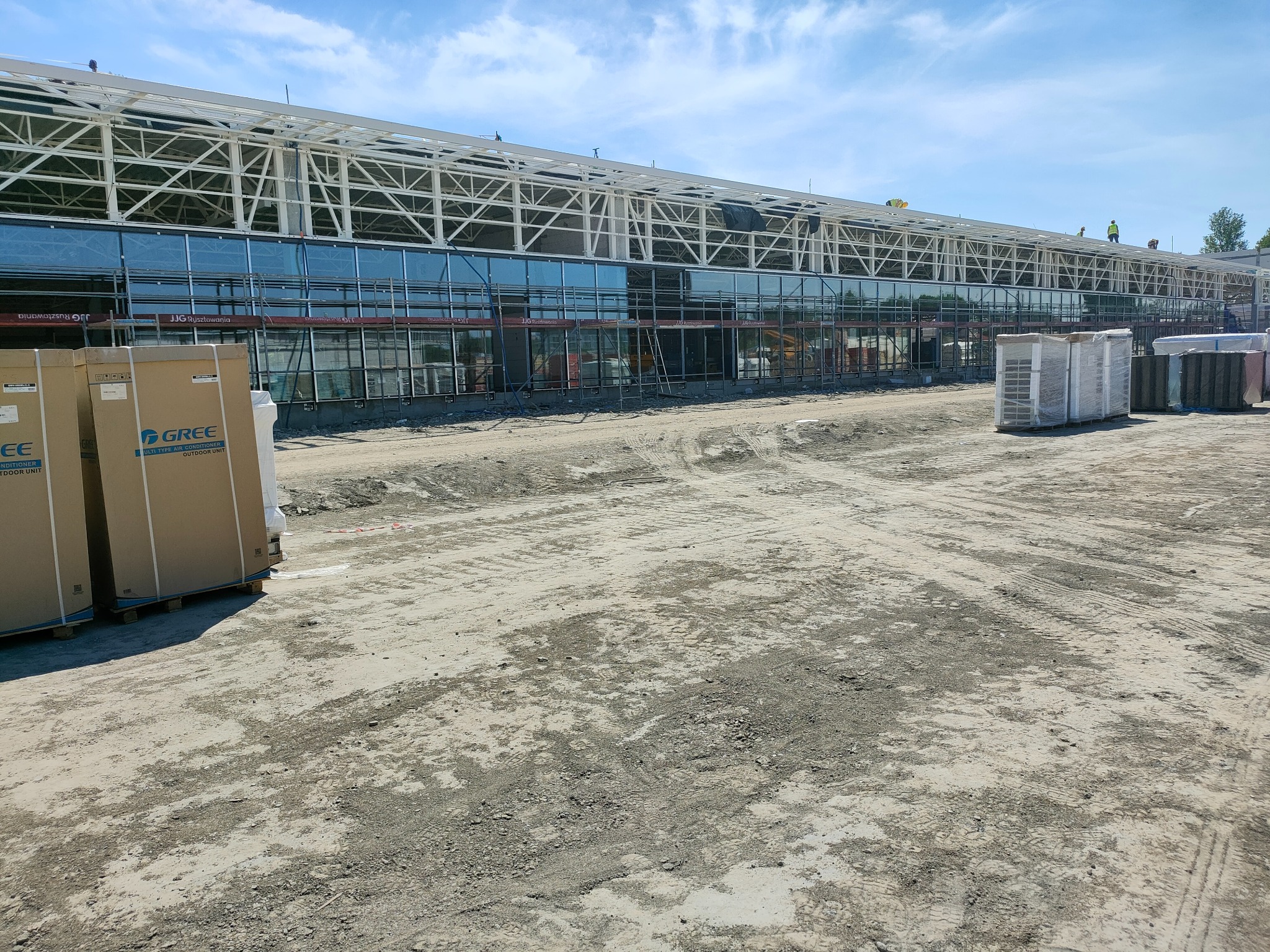 Budynek komercyjny w trakcie budowy z widocznymi rusztowaniami i pracownikami na dachu, z elementami konstrukcji stalowej i dużymi oknami fasadowymi, na pierwszym planie kartony z klimatyzatorami...