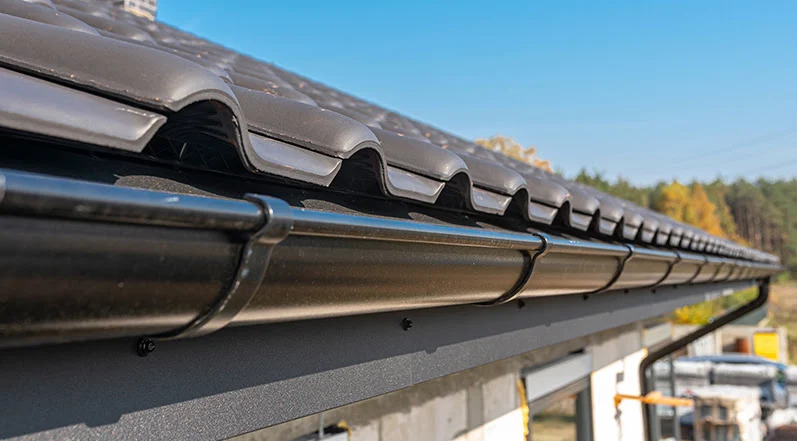 Zamontowany system rynnowy w kolorze grafitowym na dachu z dachówki ceramicznej, widok z dołu na tle błękitnego nieba i drzew.