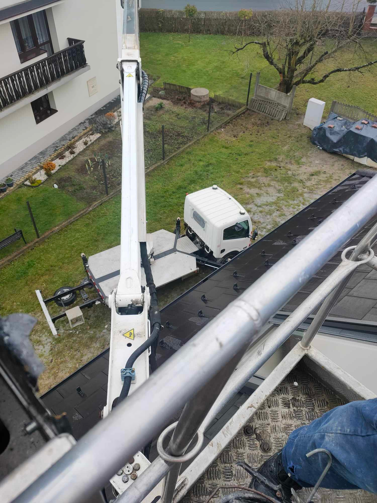 Widok z góry na podnośnik koszowy ustawiony obok budynku mieszkalnego, z widocznym fragmentem dachu, ogrodu i białej ciężarówki na posesji.