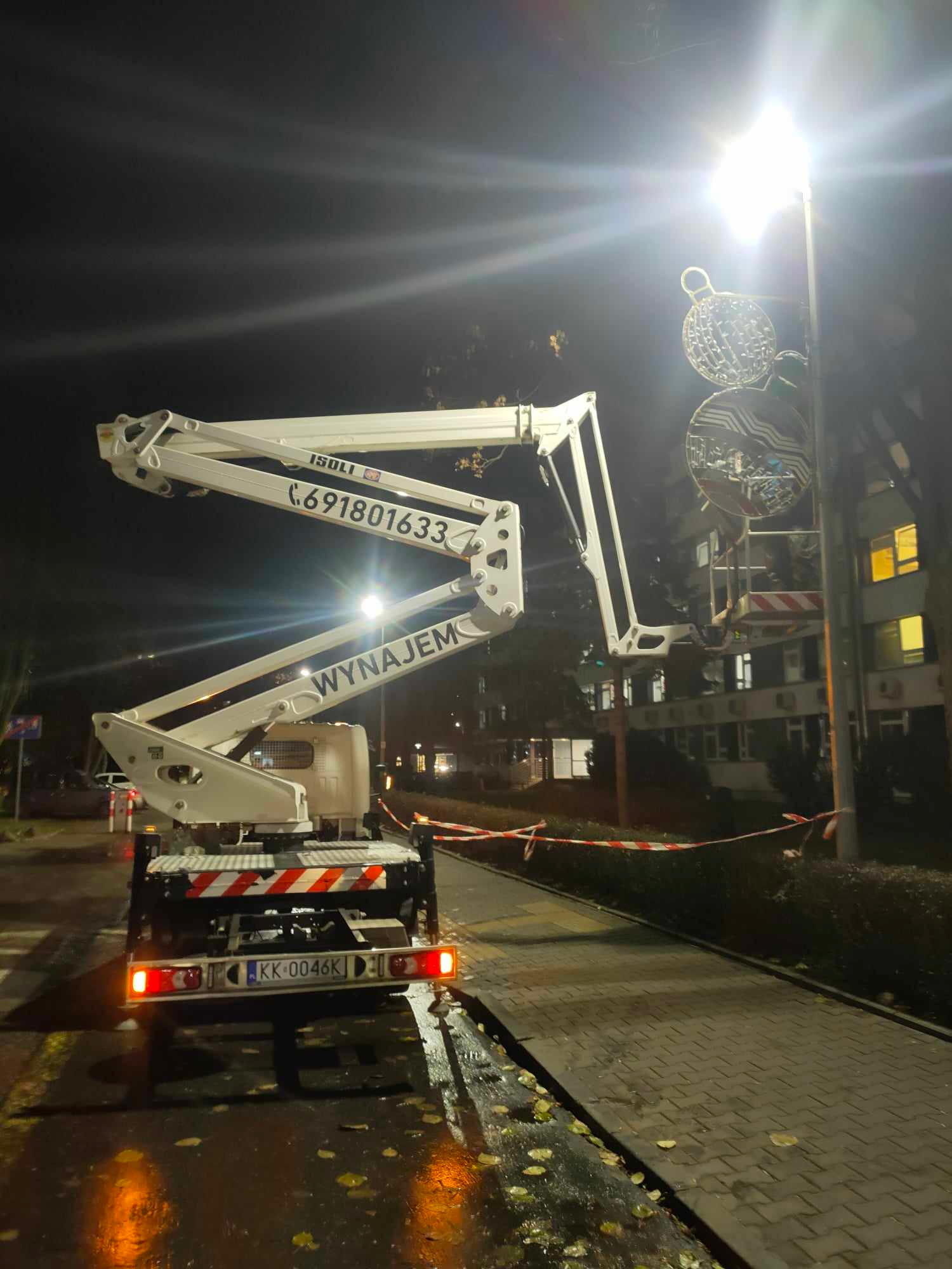 Biały podnośnik koszowy na chodniku oświetlonym latarnią, w tle budynek z oknami, na latarni zawieszone dekoracje świąteczne.