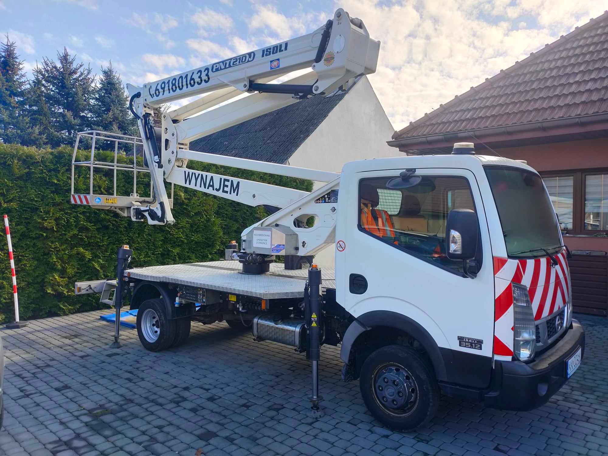 Biały podnośnik koszowy na samochodzie dostawczym, zaparkowany na brukowanym podjeździe przed budynkiem z ciemną dachówką i zielonym żywopłotem. Kosz podnośnika wysunięty, widoczny napis 'WYNAJEM'...