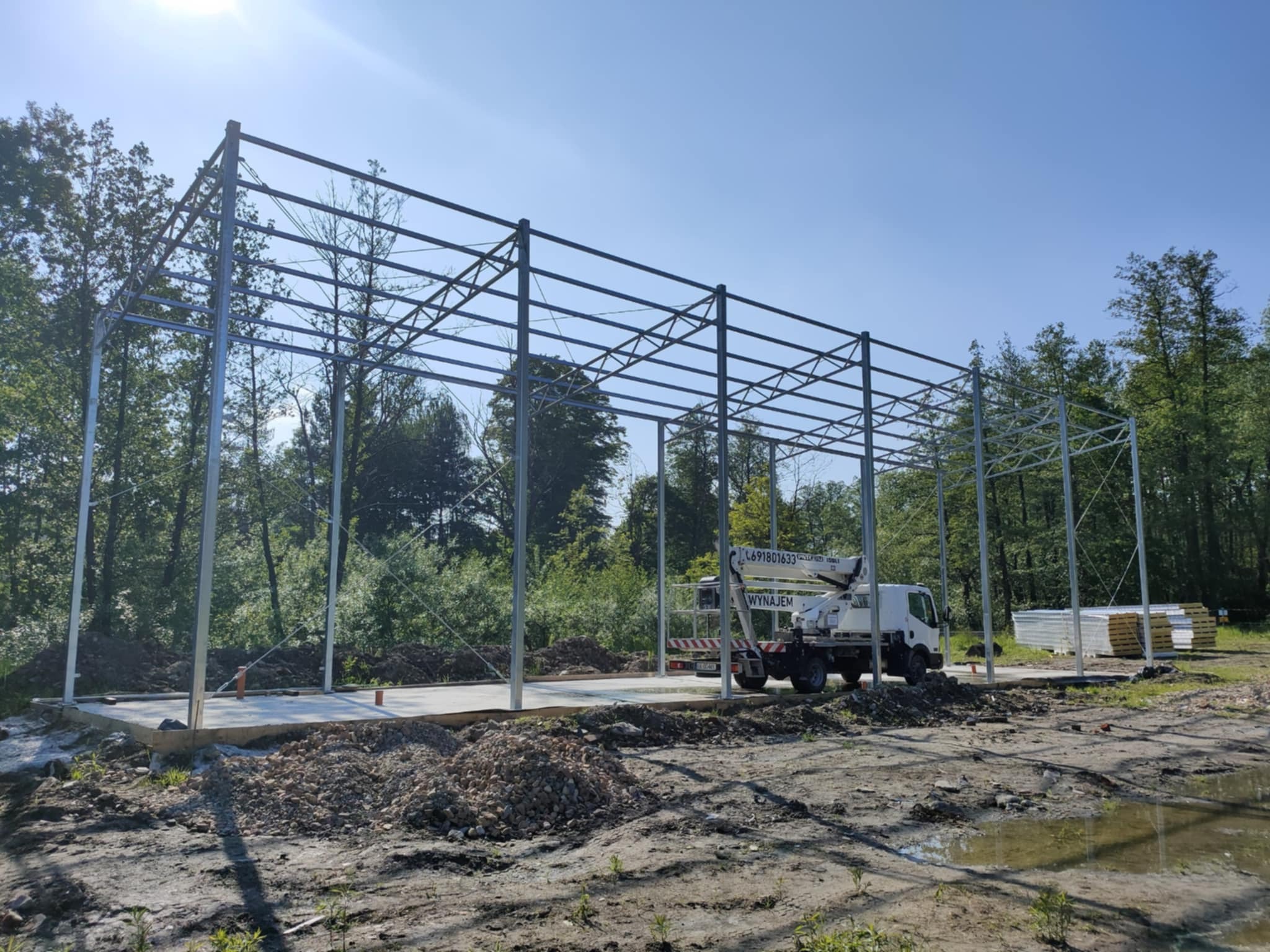 Konstrukcja stalowa hali w trakcie montażu, widoczny podnośnik koszowy, fundament betonowy, drzewa w tle i niebo. Etap budowy z bliska.