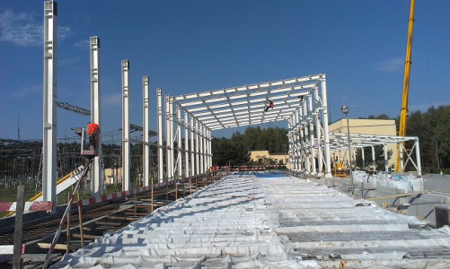 Wznoszenie konstrukcji stalowej z użyciem podnośnika koszowego i dźwigu na tle błękitnego nieba i zieleni. Widoczne białe słupy i elementy szkieletu.