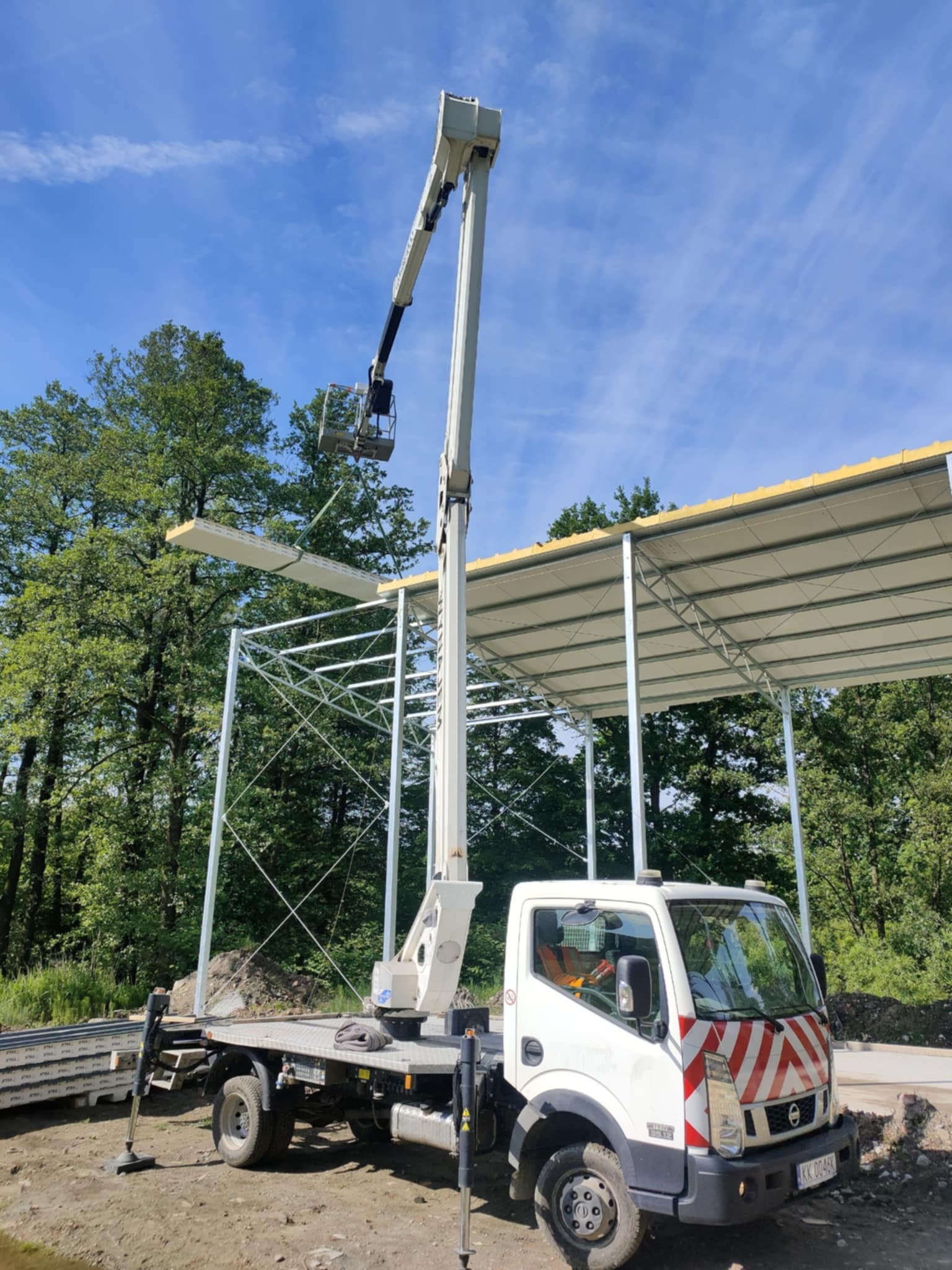 Montaż płyty warstwowej na stalowej konstrukcji z użyciem podnośnika koszowego. Widoczny fragment lasu w tle i niebieskie niebo.