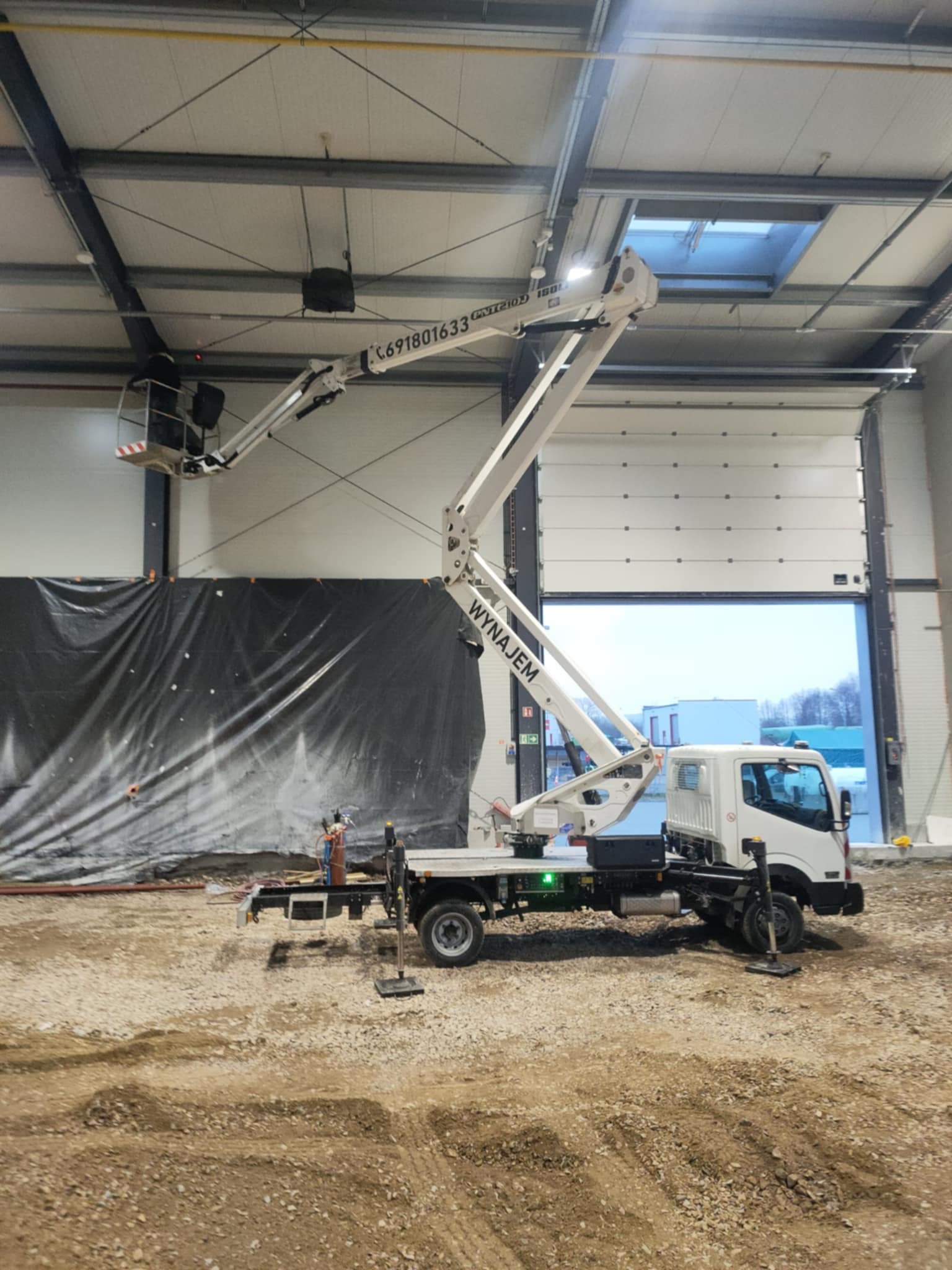 Podnośnik koszowy z pracownikiem w koszu, na tle hali z otwartą bramą. Widoczny fragment elewacji i plac budowy. Na ramieniu podnośnika numer telefonu.