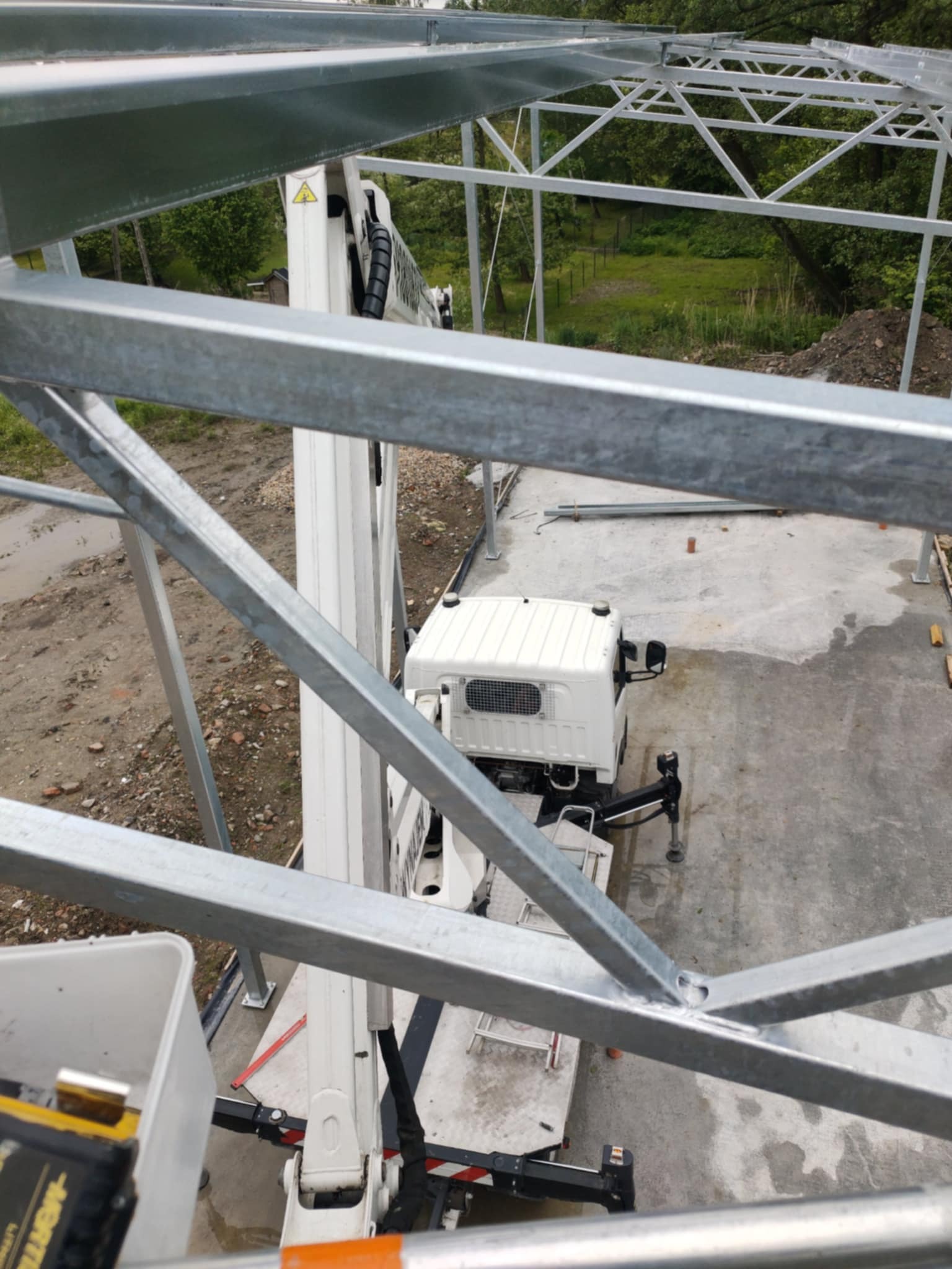 Widok z góry na konstrukcję metalową w trakcie montażu, z użyciem podnośnika koszowego. Widoczne elementy konstrukcji, podnośnik oraz otoczenie placu budowy.