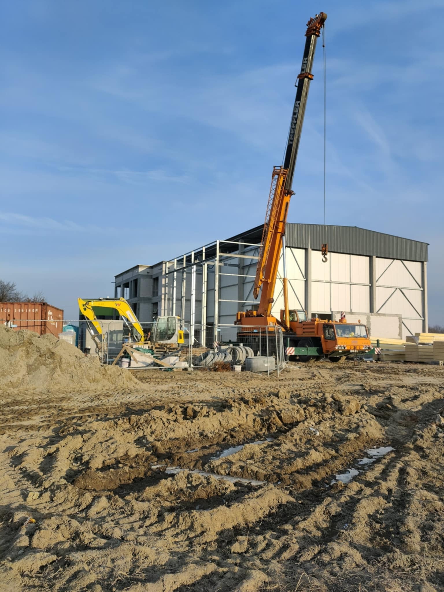 Budowa hali z płyt warstwowych w toku: widoczny dźwig Treffler, elementy konstrukcji, płyty, wykop i sprzęt budowlany. Jasne niebo w tle.