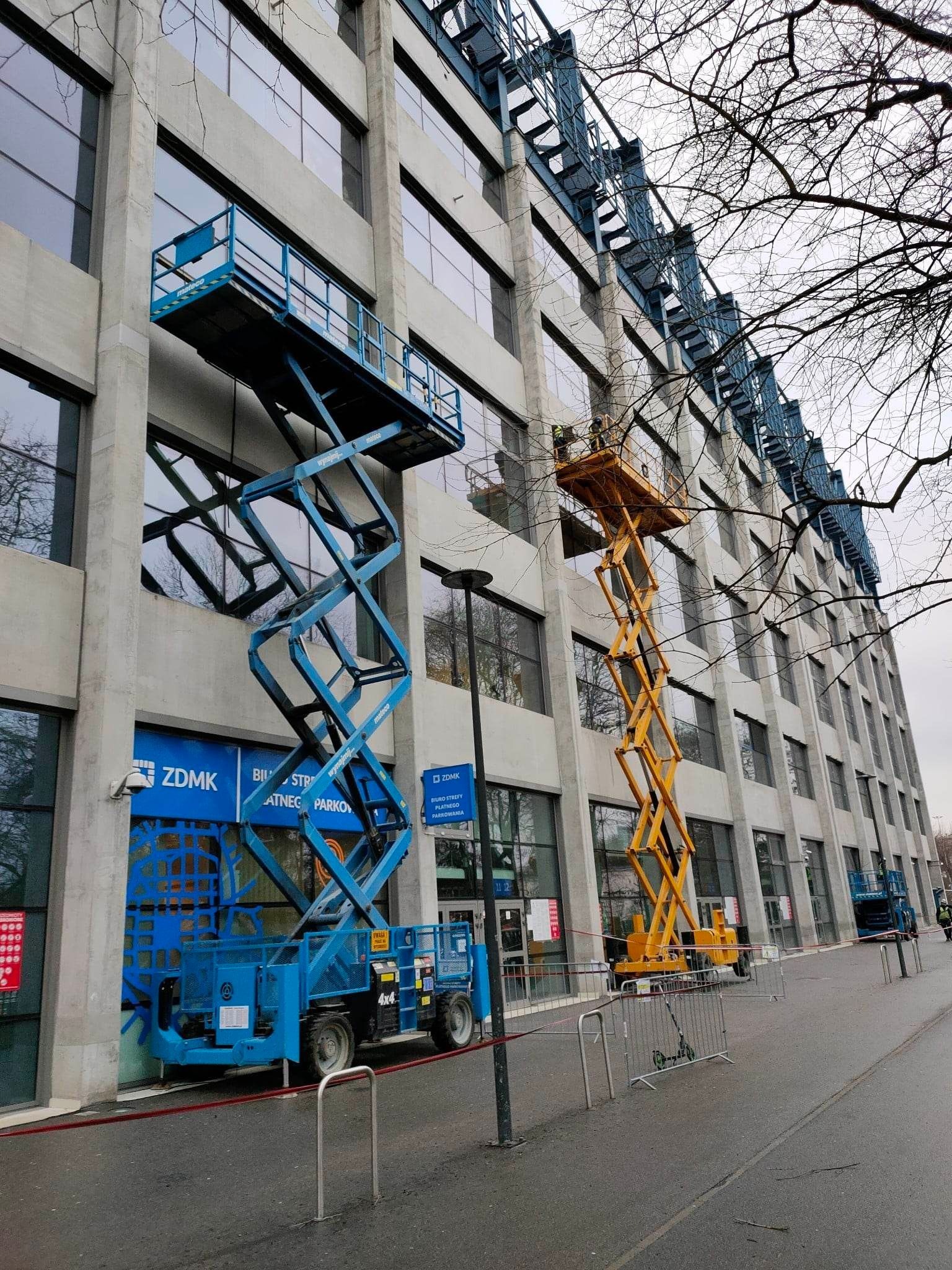 Dwa podnośniki koszowe: niebieski i żółty, ustawione przy szarym budynku z licznymi oknami. Jeden pracownik na żółtym podnośniku. Gałęzie drzew na pierwszym planie.