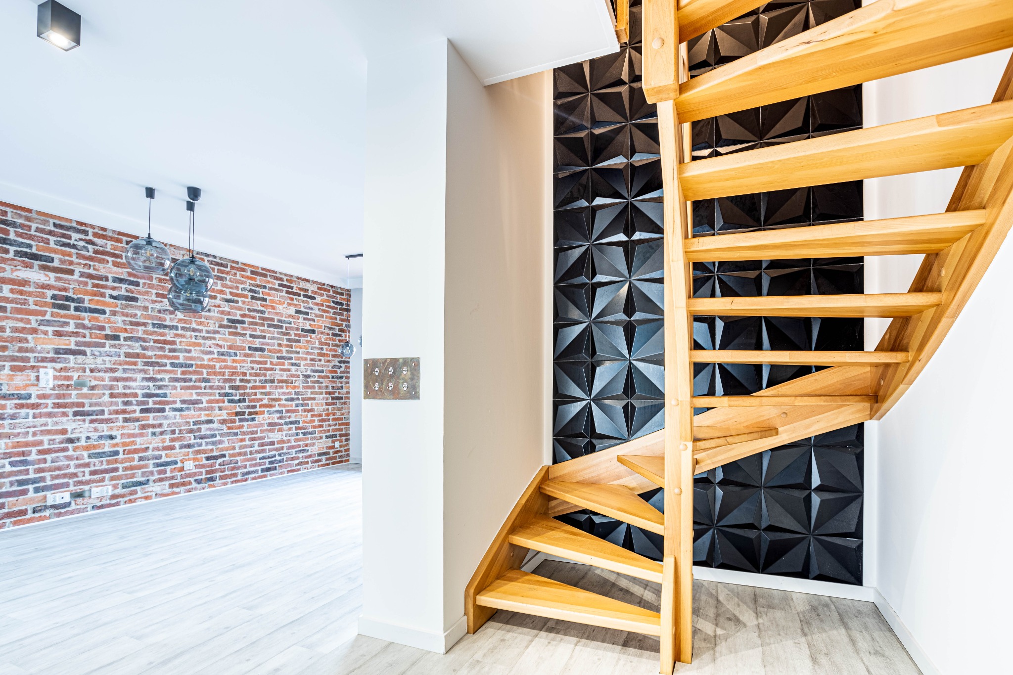 Jasne drewniane schody z ażurową konstrukcją na tle ściany z czarnymi trójwymiarowymi panelami dekoracyjnymi, w tle widoczna ceglana ściana i nowoczesne lampy wiszące.