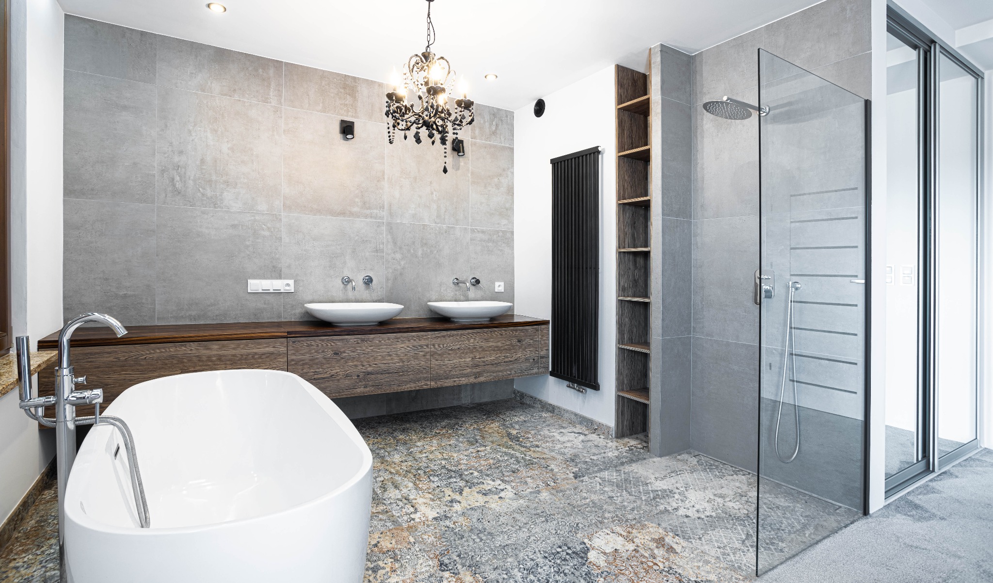Nowoczesna łazienka z wolnostojącą białą wanną, drewnianą szafką pod umywalkami i szklaną kabiną prysznicową, oświetlona ozdobnym żyrandolem.