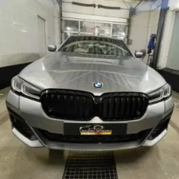 Przód srebrnego BMW serii 5 z czarnym grillem, po detailingu, widoczny znak firmowy na dole zderzaka, kratka odpływowa na podłodze, brama garażowa w tle.