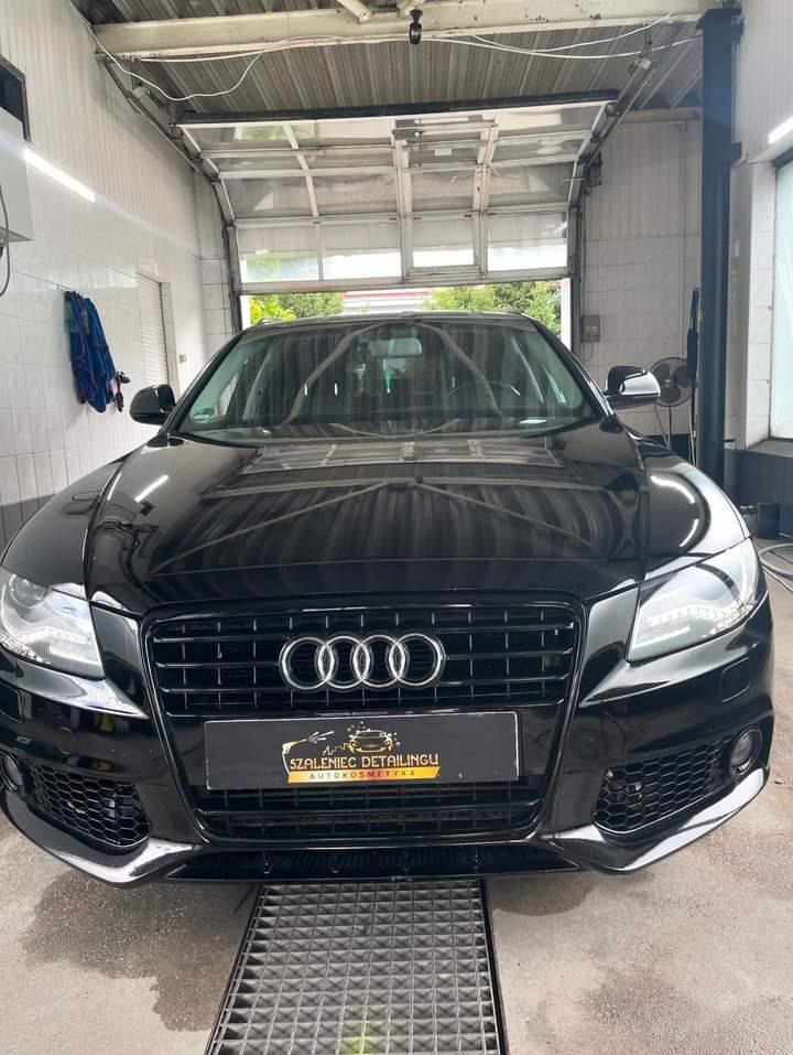 Błyszczący, czarny samochód Audi po detailingu, zaparkowany w jasnym garażu z kratką odpływową w podłodze i widocznym logo firmy detailingu na przednim grillu.