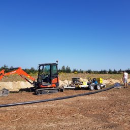 KOKODRIL JANUSZ BAŁUT - Pomarańczowa mini koparka Kubota KX030-4 na placu budowy obok przyczepy z agregatem prądotwórczym i długiego odcinka czarnej rury, w tle skarpa z piasku i pracownik w białej koszulce.
