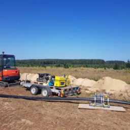 Pomarańczowa mini koparka Kubota, przyczepa z agregatem i spawarką do rur PE, oraz długa czarna rura PE na ziemi, w tle las i błękitne niebo.