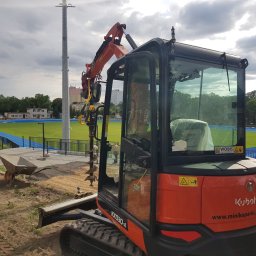 KOKODRIL JANUSZ BAŁUT - Pomarańczowa minikoparka Kubota KX030-4 z wiertłem do gruntu, pracująca na terenie budowy obok stadionu lekkoatletycznego z niebieską bieżnią, widoczny pracownik z taczką i narzędziami.