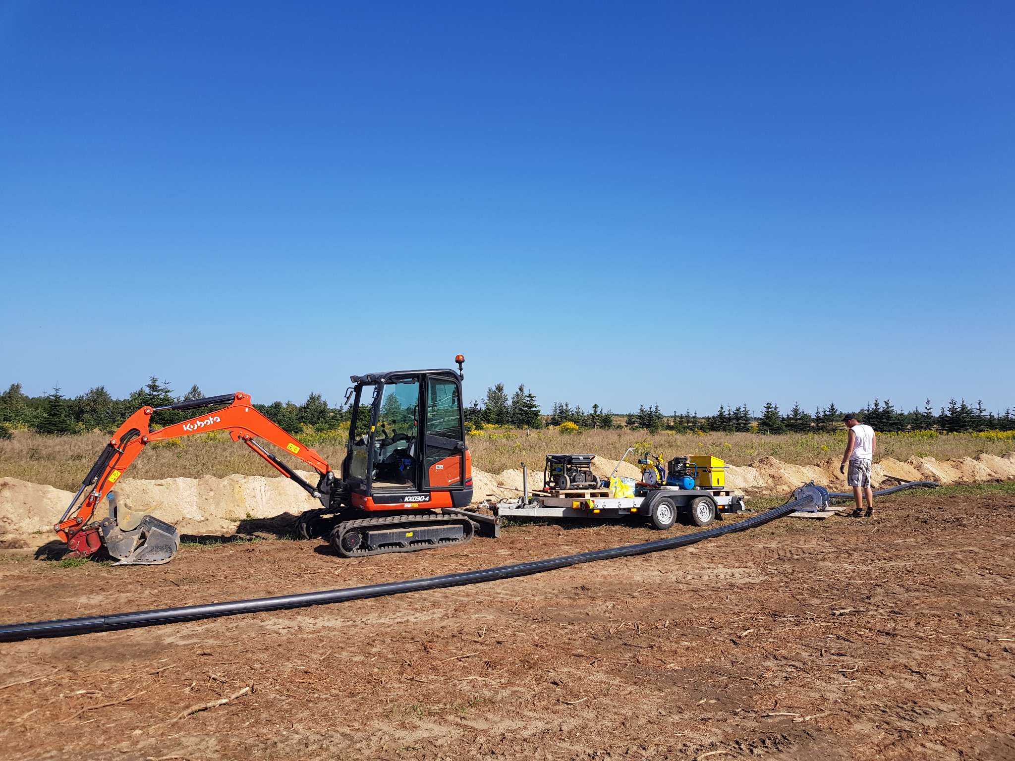 Pomarańczowa mini koparka Kubota KX030-4 na placu budowy obok przyczepy z agregatem prądotwórczym i długiego odcinka czarnej rury, w tle skarpa z piasku i pracownik w białej koszulce.