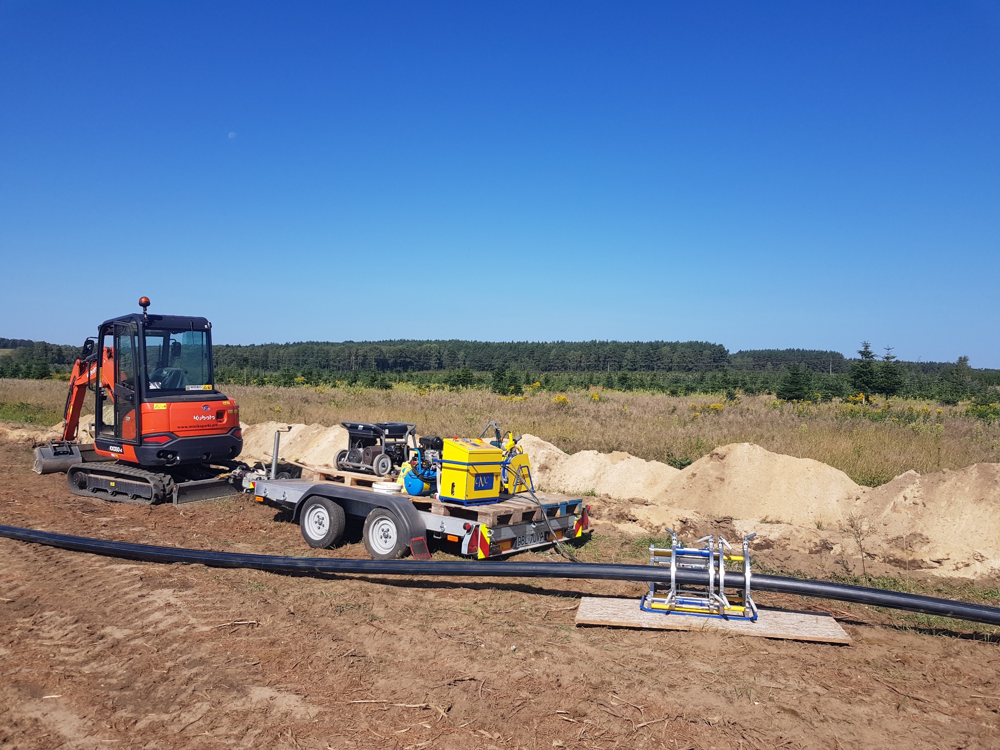 Pomarańczowa mini koparka Kubota, przyczepa z agregatem i spawarką do rur PE, oraz długa czarna rura PE na ziemi, w tle las i błękitne niebo.