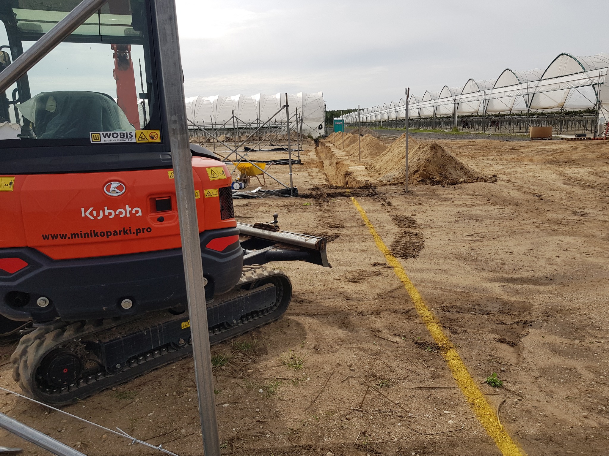 Pomarańczowa minikoparka Kubota w Nowogrodźcu, widoczna na tle wykopanego rowu i konstrukcji szklarni, z żółtą linią wyznaczającą obszar robót.
