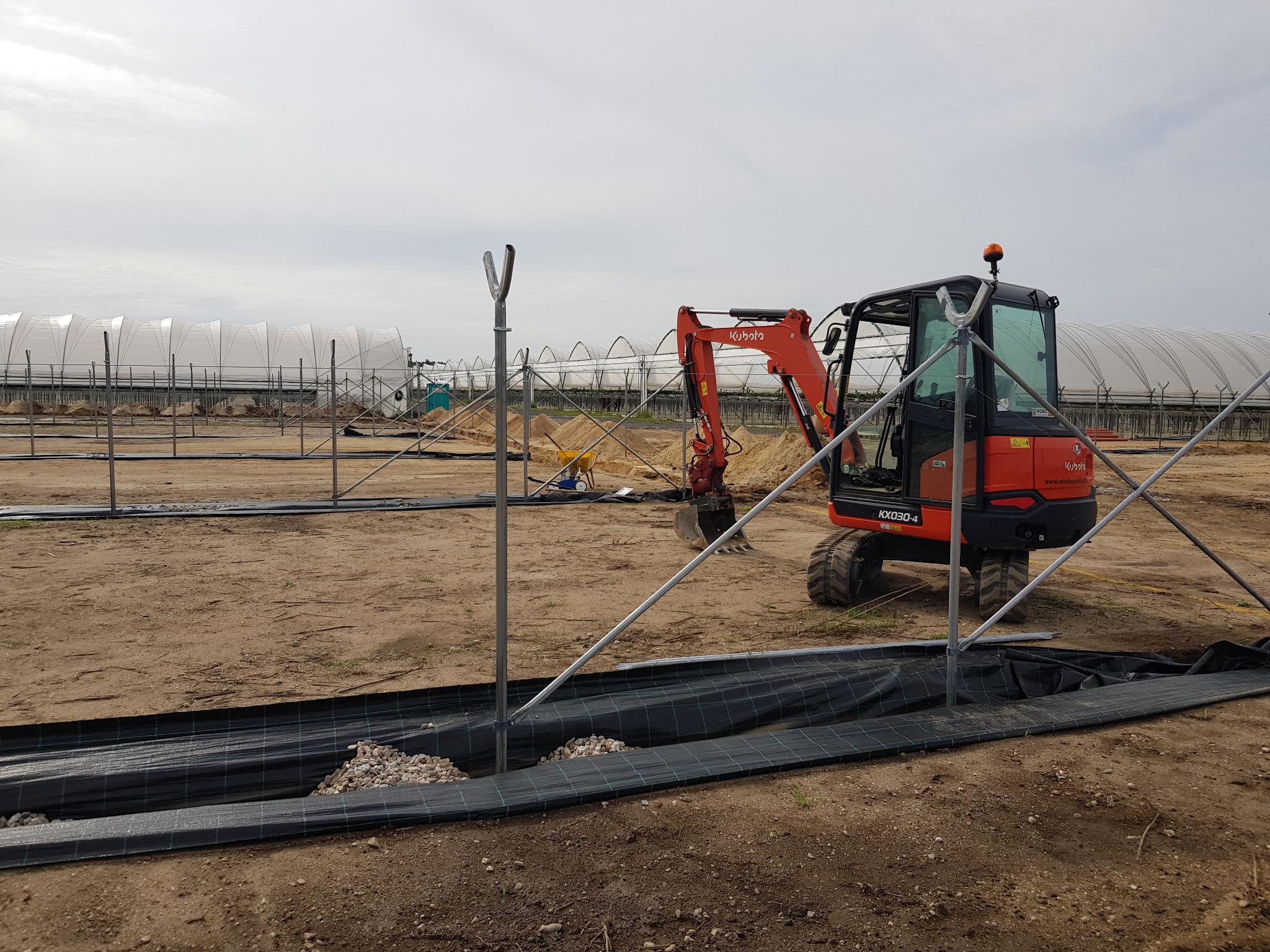Mała koparka Kubota KX030-4 na placu budowy z przygotowanym podłożem z czarnej geowłókniny i kamieniami, w tle szkielety tuneli foliowych.