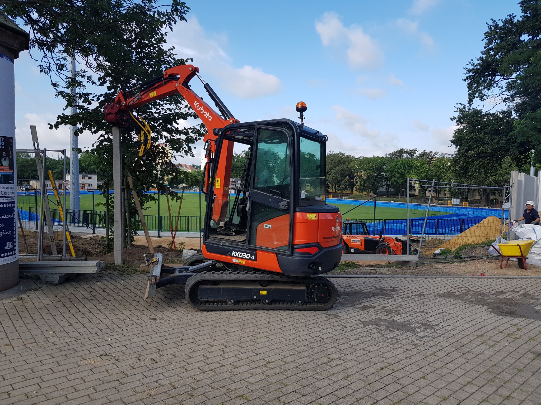 Pomarańczowa mini koparka Kubota KX030-4 na brukowanej powierzchni, w tle ogrodzone boisko sportowe z niebieską nawierzchnią i inna maszyna budowlana, widoczny pracownik w kasku.