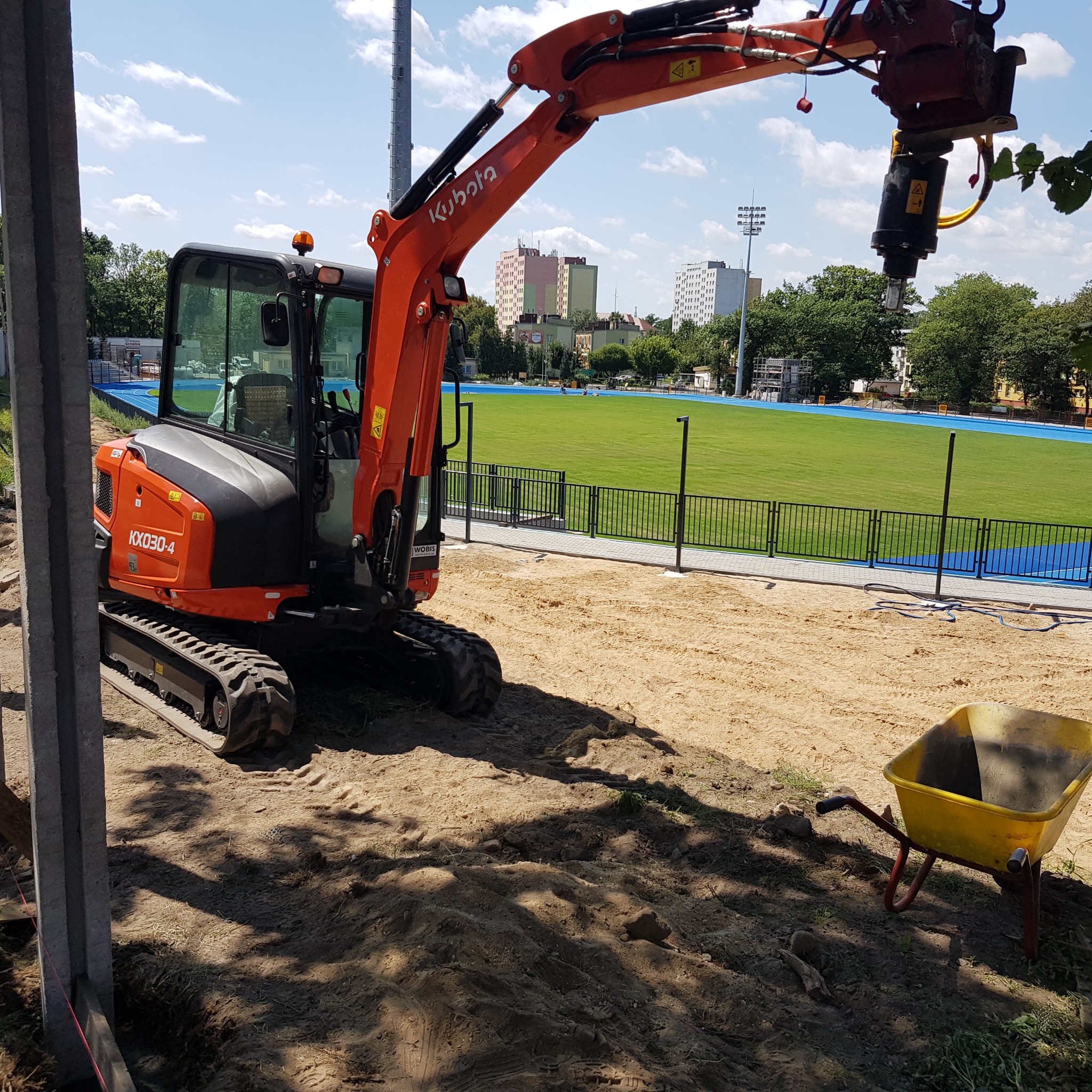 Pomarańczowa minikoparka Kubota z wiertnicą hydrauliczną na placu budowy stadionu, obok żółta taczka, w tle budynki miejskie i oświetlenie stadionu.