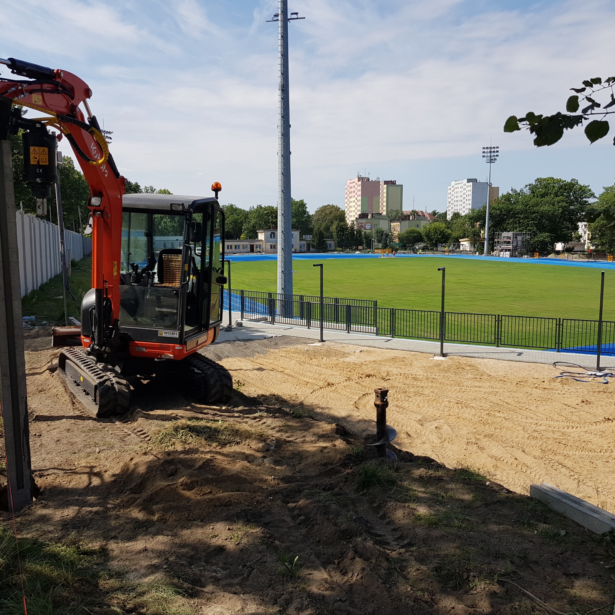 Pomarańczowa mini koparka Kubota z wiertłem do gruntu na placu budowy, w tle boisko sportowe z niebieską bieżnią i budynki mieszkalne.