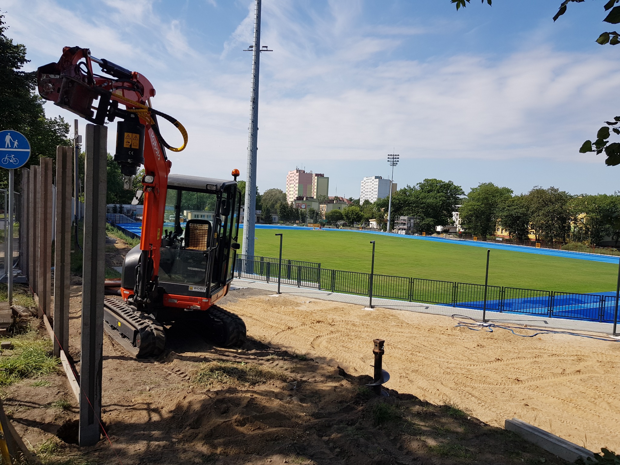 Pomarańczowa mini-koparka z wiertnicą hydrauliczną wbija pale ogrodzenia na placu budowy stadionu lekkoatletycznego z zieloną murawą i niebieską bieżnią.