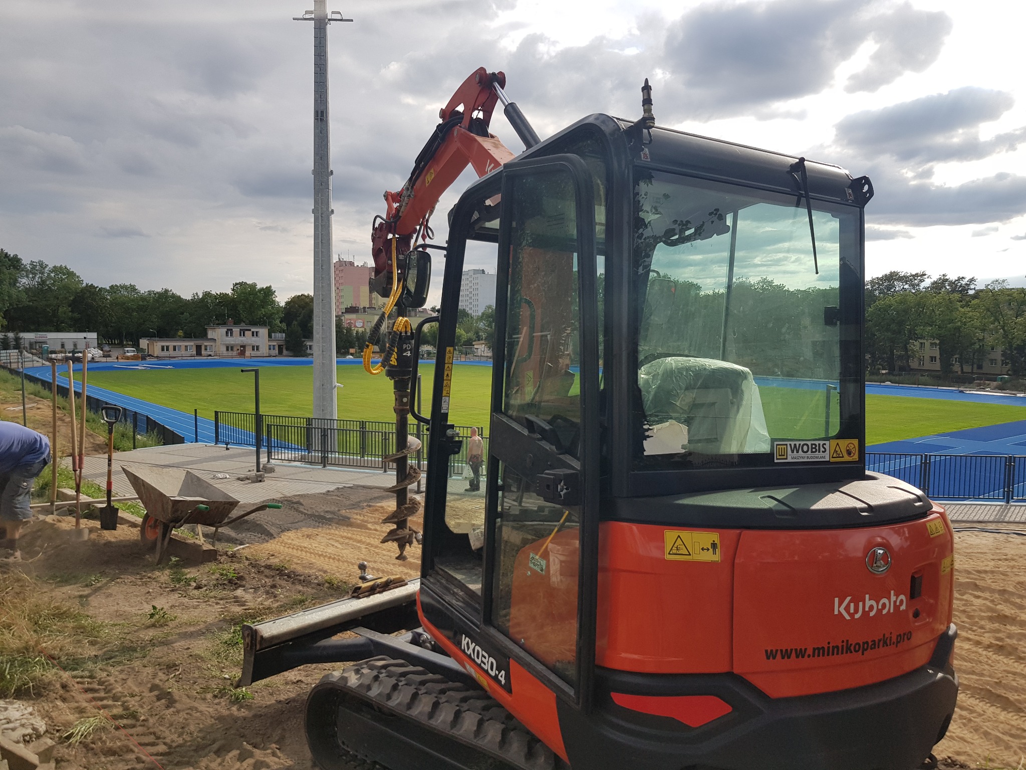 Pomarańczowa minikoparka Kubota KX030-4 z wiertłem do gruntu, pracująca na terenie budowy obok stadionu lekkoatletycznego z niebieską bieżnią, widoczny pracownik z taczką i narzędziami.