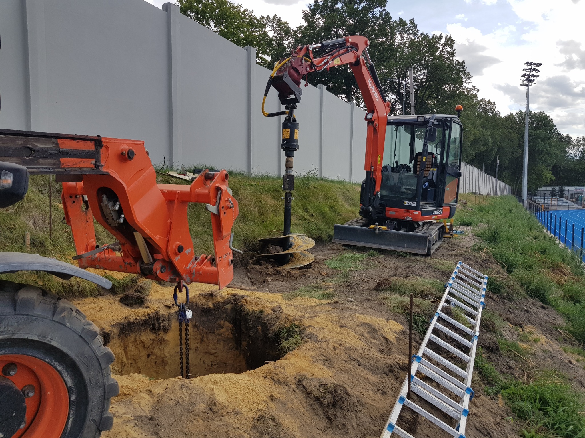 Pomarańczowa koparka z wiertłem świdrowym wierci otwór w ziemi obok wykopu, w tle stadion z niebieską bieżnią i oświetleniem.