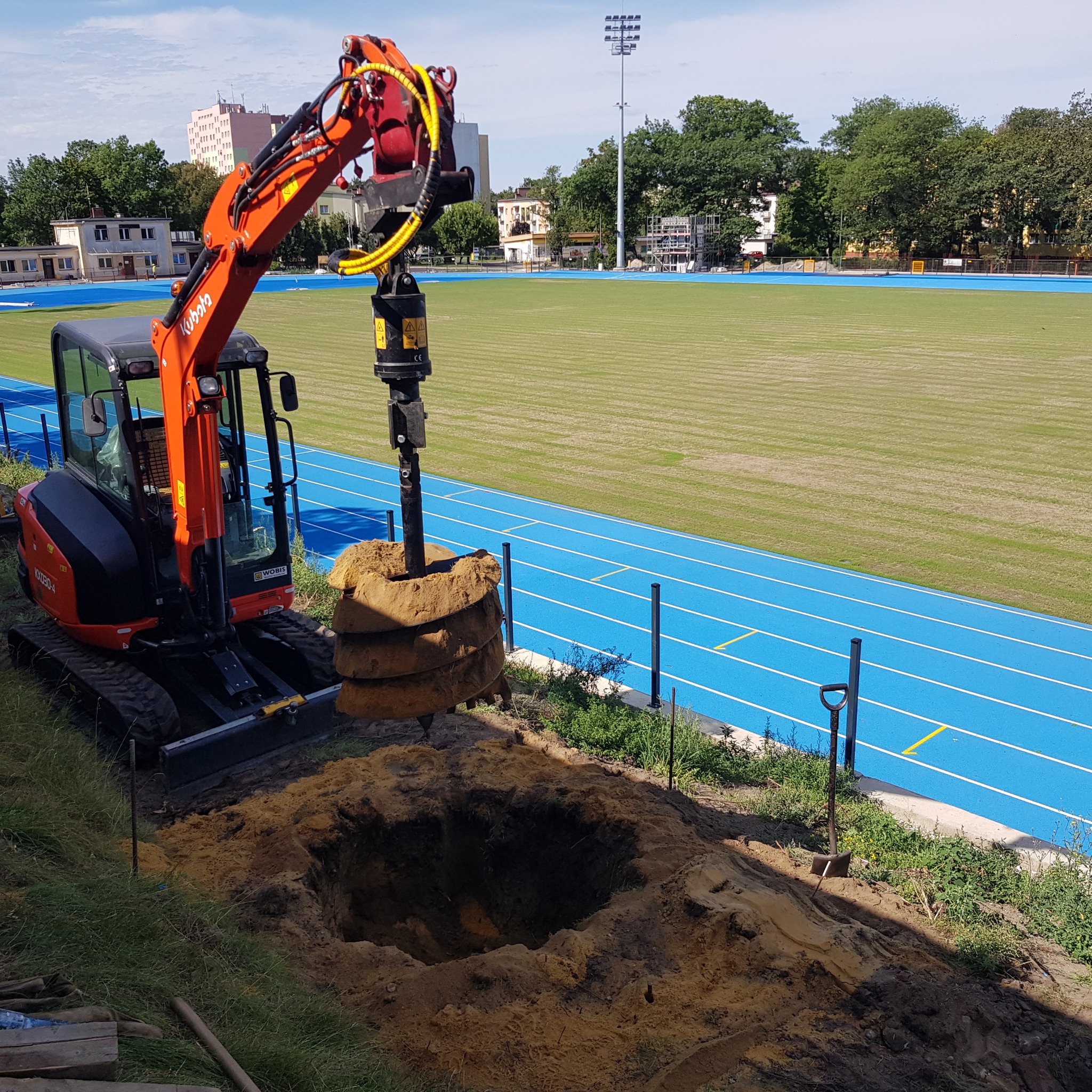 Pomarańczowa minikoparka Kubota z wiertnicą wydobywa rdzeń ziemi z wywierconego otworu na tle niebieskiej bieżni tartanowej i zielonego boiska.