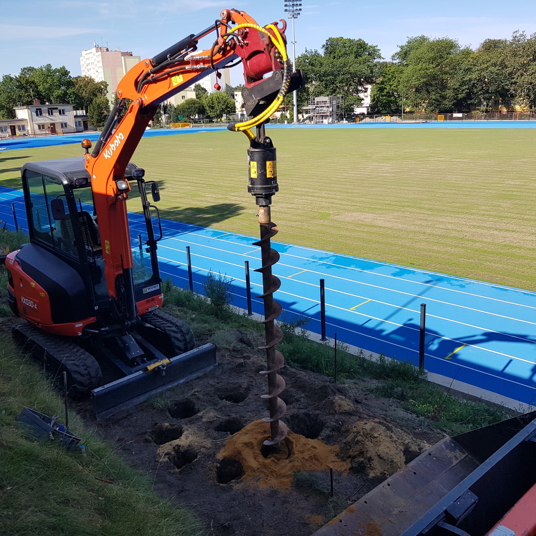 Pomarańczowa minikoparka Kubota z wiertnicą hydrauliczną wykonuje otwory w ziemi, widoczne liczne wywiercone dołki, w tle błękitna bieżnia stadionu.
