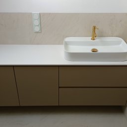 POLI IMAGER - Nowoczesna łazienka z beżową szafką pod umywalką, białą prostokątną umywalką nablatową i złotą baterią. Na ścianach płytki imitujące marmur. Widoczny włącznik światła z trzema okrągłymi przyciskami.