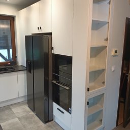 POLI IMAGER - Nowoczesna, biała zabudowa kuchenna z czarnym sprzętem AGD: lodówka side-by-side, piekarnik i szklana witryna. Minimalistyczne uchwyty. Szare płytki podłogowe.