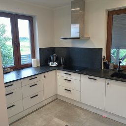 POLI IMAGER - Nowoczesna kuchnia z białymi szafkami i czarnymi blatami, okap ze stali nierdzewnej, dwa okna z widokiem na zieleń. Minimalistyczny design z akcentami kolorystycznymi.